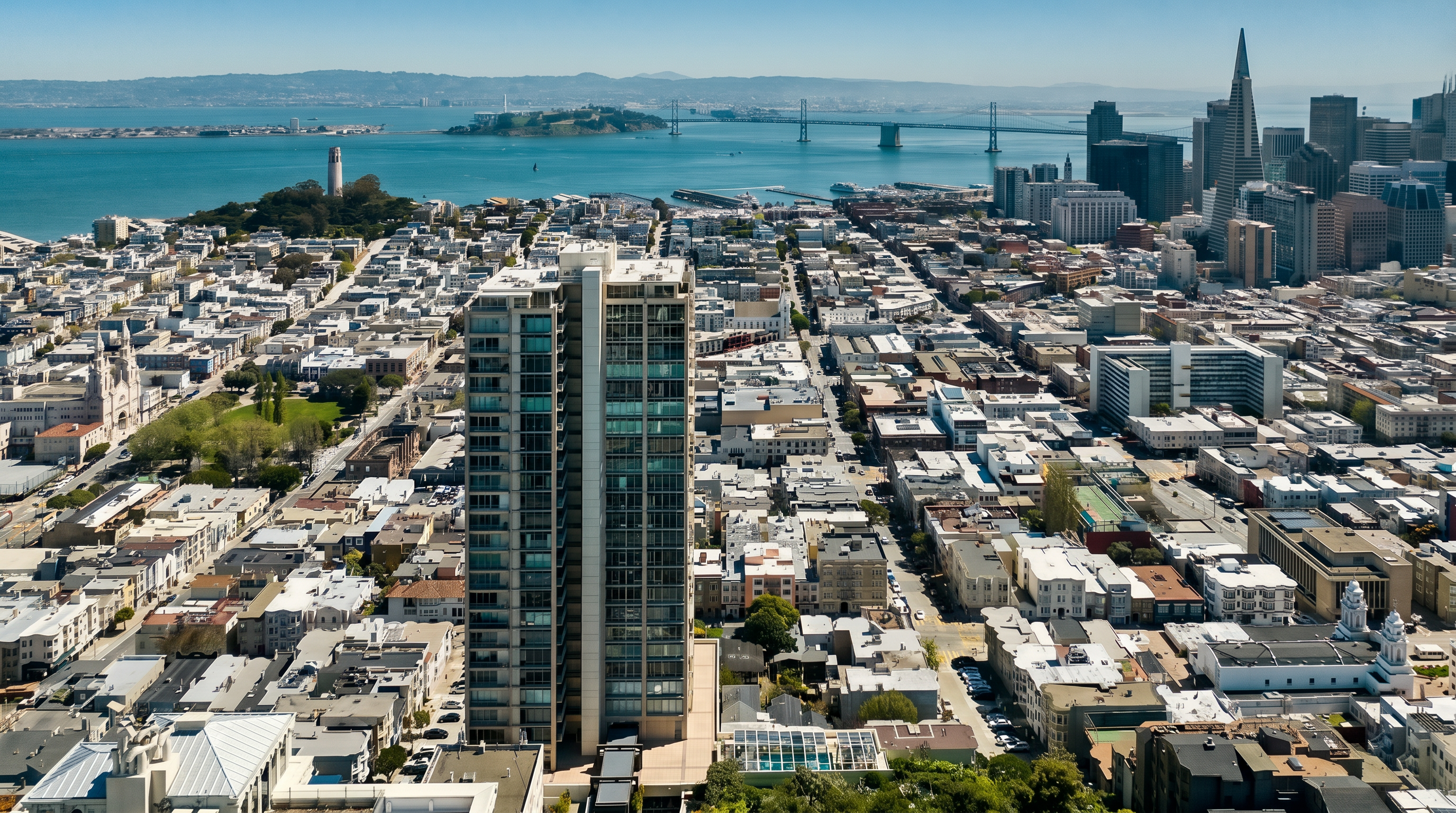 This high-altitude aerial view captures a prominent residential high-rise tower in San Francisco, showcasing its central position within the dense urban grid. The perspective highlights the building's relationship to the surrounding neighborhood, the iconic Coit Tower, and the expansive San Francisco Bay with the Bay Bridge in the distance. The scene provides a comprehensive look at the city's architectural density and coastal geography on a clear, sunny day.