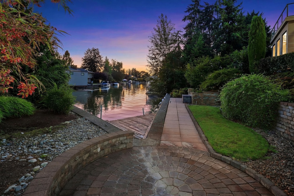 This serene waterfront backyard features a circular paver patio that transitions into a stone-lined walkway leading toward a private wooden dock. Lush landscaping, including mature trees and manicured shrubs, frames the tranquil canal view during a vibrant sunset. The perspective is low and wide, capturing the peaceful transition from the hardscaped garden area to the water's edge.