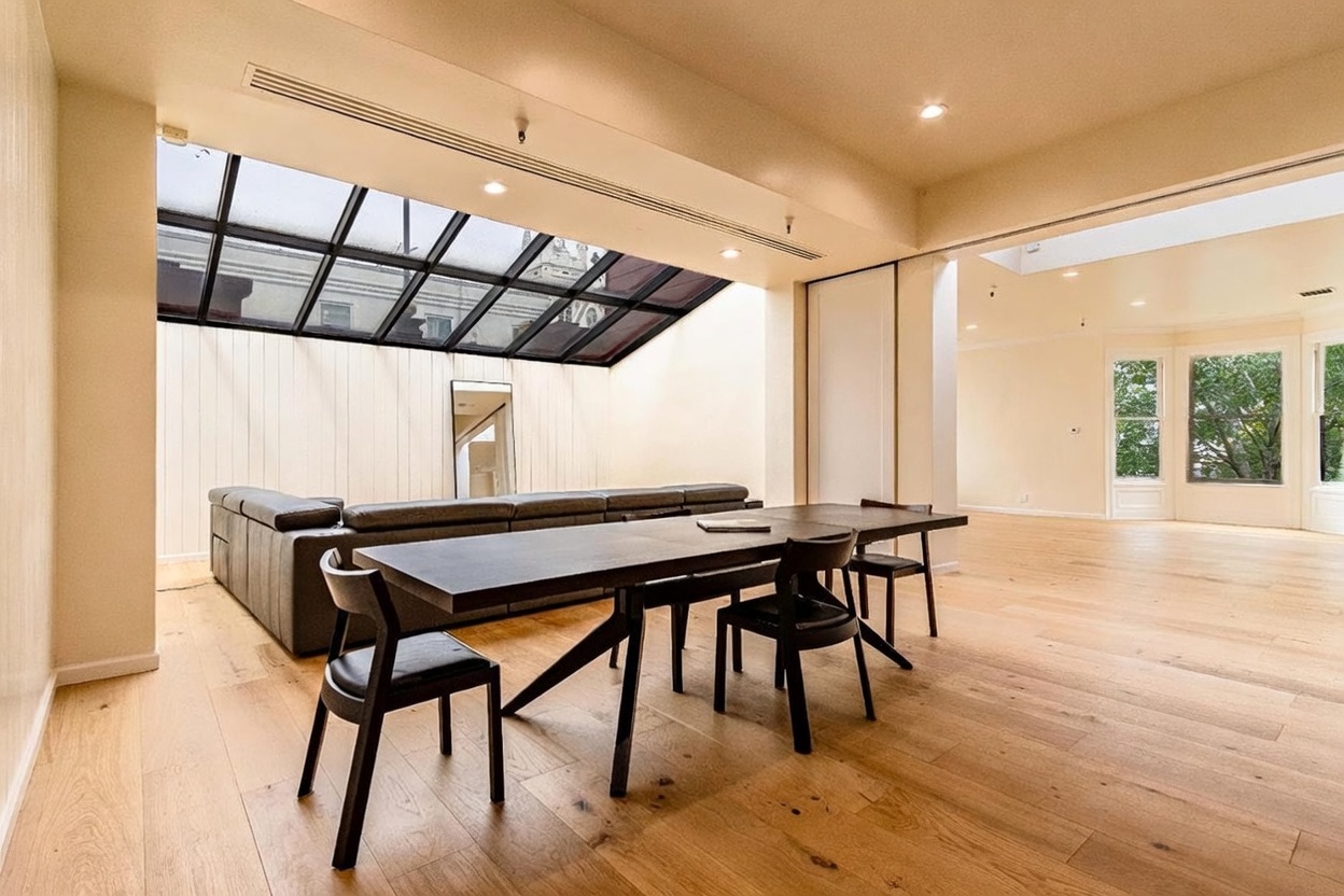 This spacious, open-concept living area features a large, dark-toned sectional sofa positioned beneath a dramatic, sloped glass skylight that floods the room with natural light. A long, dark wood dining table sits in the foreground, creating a versatile space that flows seamlessly into an adjacent, bright room with large windows. The interior is characterized by light wood flooring, neutral walls, and a clean, contemporary aesthetic.