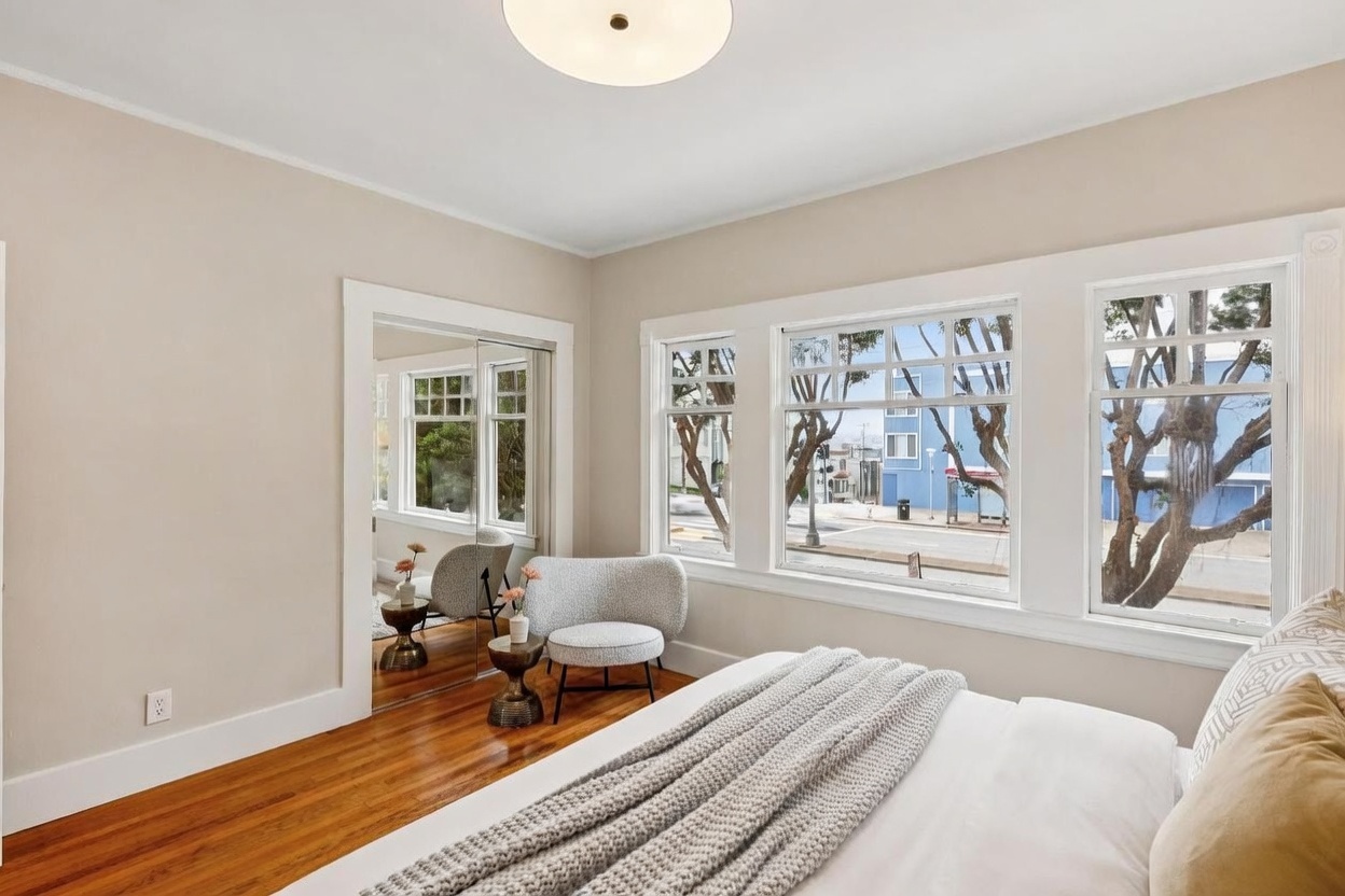 This bright and airy bedroom features warm wood flooring and neutral-toned walls, creating a welcoming atmosphere. A cozy armchair sits beside a small side table near a set of mirrored closet doors, while large windows provide ample natural light and a view of the street outside. The bed is dressed in a textured gray throw blanket, emphasizing a comfortable and inviting aesthetic.