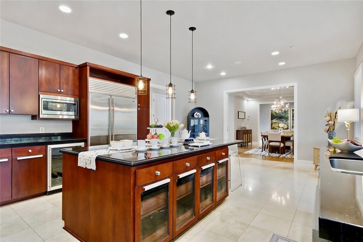 This spacious kitchen features dark wood cabinetry, a large central island with a dark stone countertop, and stainless steel appliances. Three pendant lights hang above the island, which is set with dishware and decorative items, while the adjacent dining area is visible through a wide doorway. The room is finished with light-colored tile flooring and recessed lighting, creating a bright and inviting atmosphere.