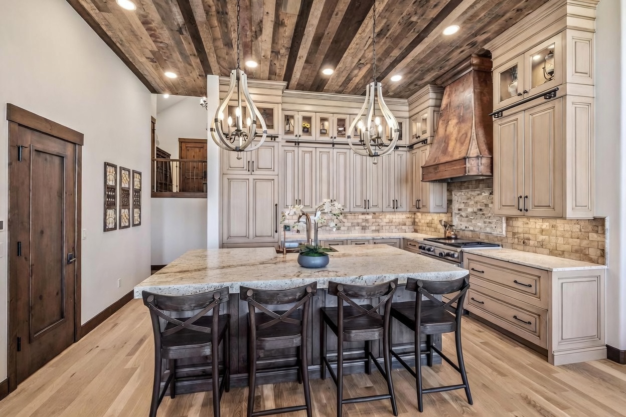 This spacious kitchen features a large central island with seating for four, topped with a light-toned stone surface. The room is defined by its rustic wood-plank ceiling, custom cabinetry in a neutral finish, and a prominent copper-toned range hood above the stove. The space is illuminated by two decorative pendant lights and recessed lighting, creating a warm and inviting atmosphere.