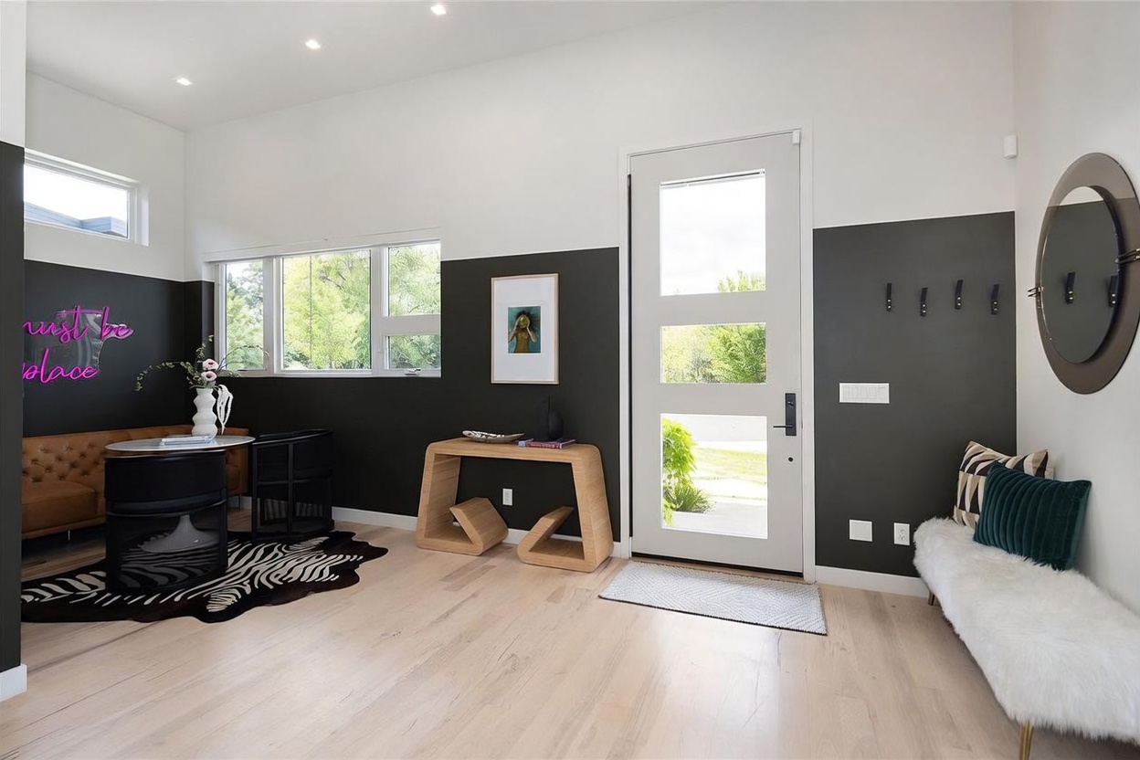 This modern entryway features a striking two-tone wall design with a dark charcoal lower section and crisp white upper walls. The space includes a unique sculptural wooden console table, a white front door with glass panels, and a plush white bench paired with a circular mirror. A zebra-print rug adds a bold, contemporary touch to the light wood flooring, creating a stylish and welcoming first impression.