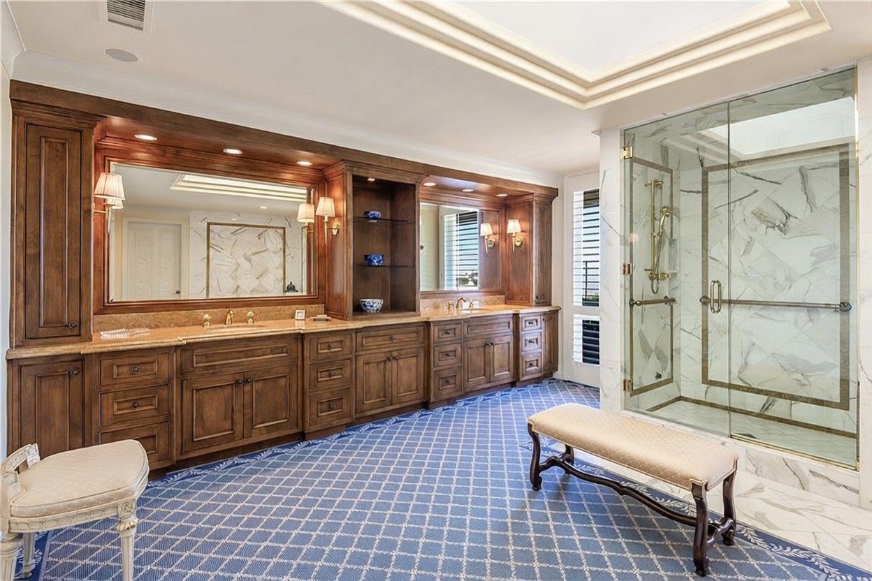 This spacious primary bathroom features a long, dark wood vanity with dual sinks and extensive cabinetry, complemented by a large wall-to-wall mirror. To the right, a glass-enclosed shower with patterned stone walls adds a luxurious touch, while a patterned blue carpet covers the floor. Two elegant benches are positioned in the room, creating a sophisticated and functional space.