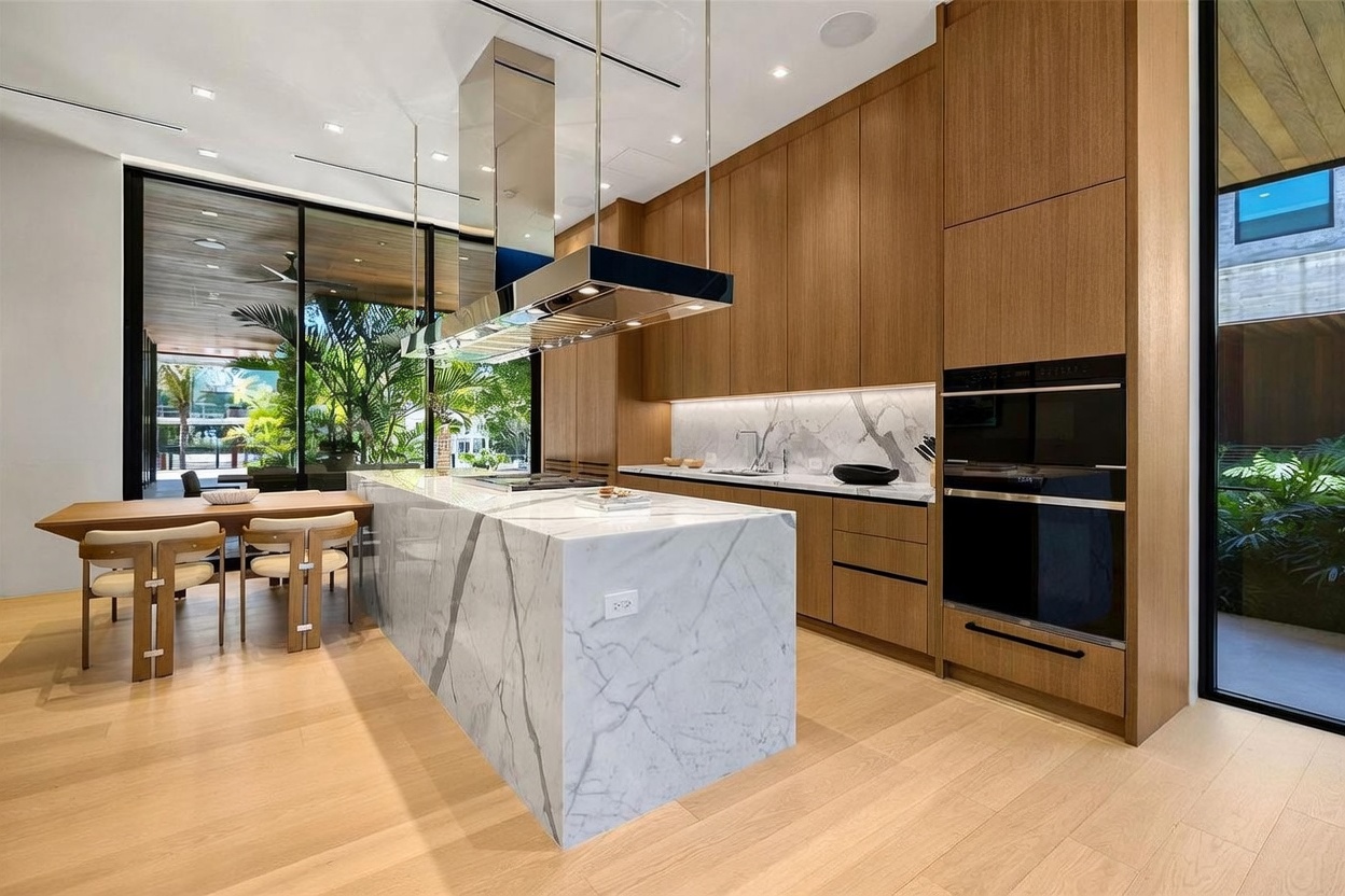 This modern, open-concept kitchen features a large central island with a stone waterfall edge and integrated cooktop, complemented by light wood cabinetry and a sleek stainless steel range hood. The space is bathed in natural light from floor-to-ceiling glass walls that provide a seamless transition to the lush outdoor greenery. A dining table is attached to the end of the island, creating a cohesive and functional layout for both cooking and entertaining.