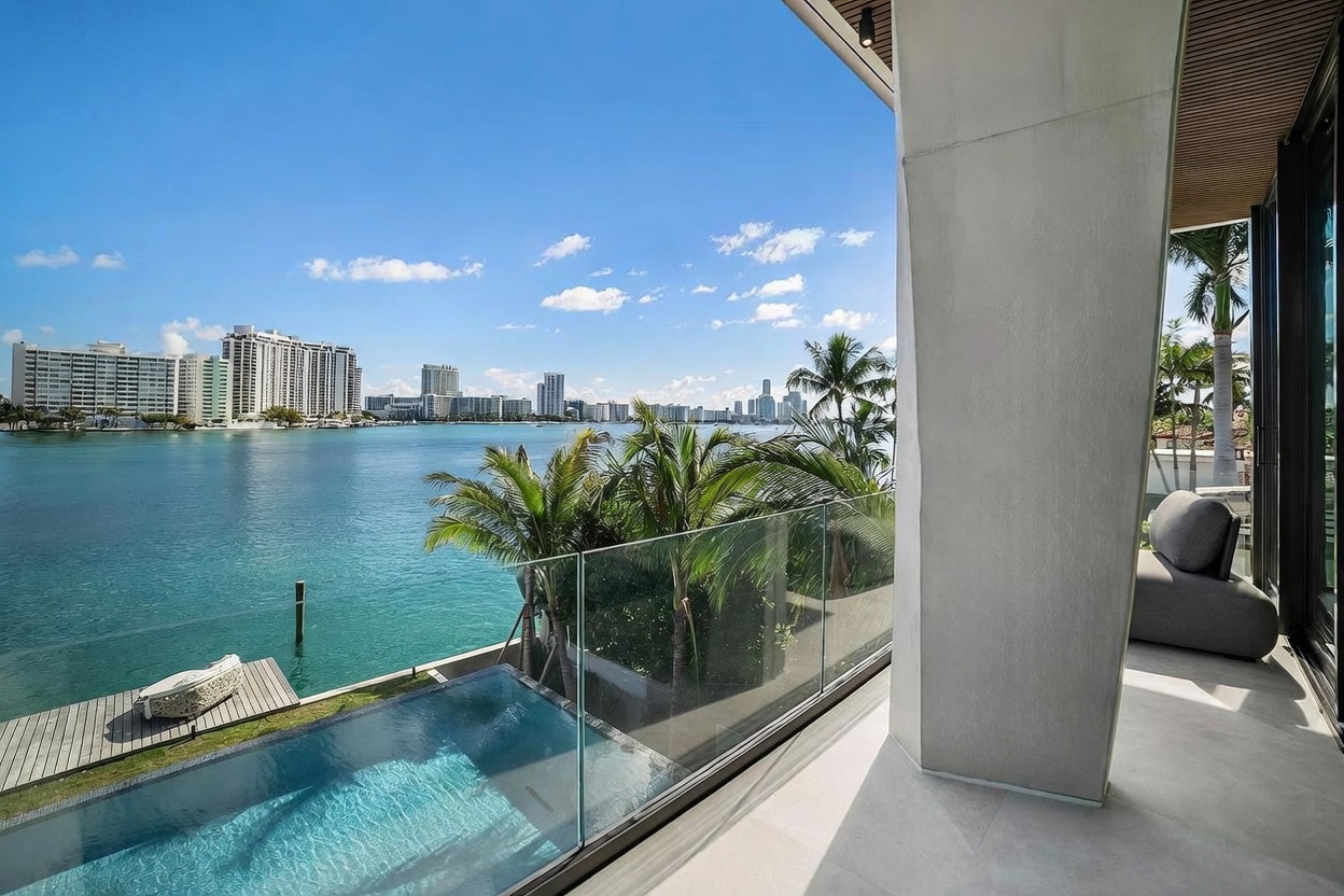 This luxurious balcony offers a panoramic waterfront view, featuring a sleek glass railing that overlooks a private infinity-edge pool and a wooden dock. The space is furnished with a comfortable grey lounge chair and is sheltered by a modern concrete structure with a slatted wood ceiling. The bright, sunny atmosphere highlights the serene coastal setting and the expansive cityscape across the water.