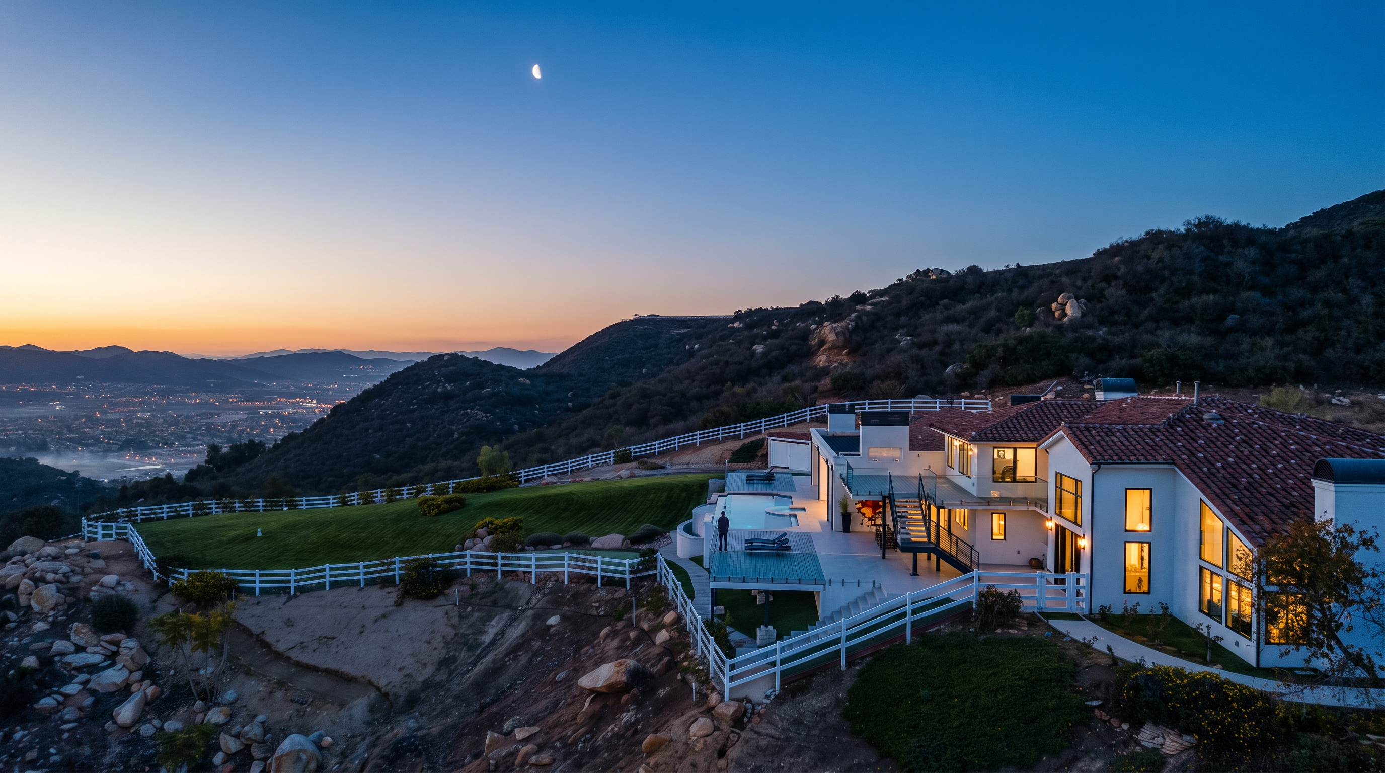 This stunning aerial perspective captures a luxury hillside estate at dusk, featuring a sprawling multi-level Mediterranean-style home with a tile roof and a private pool deck. The property boasts extensive landscaped grounds enclosed by white fencing, set against a dramatic backdrop of rolling hills and panoramic valley views under a twilight sky. This unique positioning provides an exceptional sense of privacy and scenic beauty, making it a standout luxury residence.