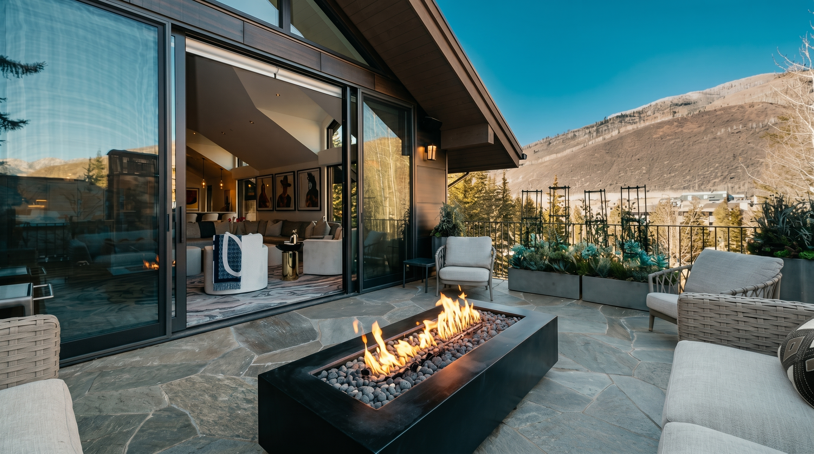 This expansive mountain-view terrace features a modern rectangular fire pit as its centerpiece, surrounded by comfortable outdoor lounge seating. Large glass sliding doors open to reveal a bright, contemporary living area, creating a seamless transition between indoor and outdoor living spaces. The stone-paved patio is framed by a metal railing and elevated planters, offering a clear view of the surrounding landscape and mountain range.