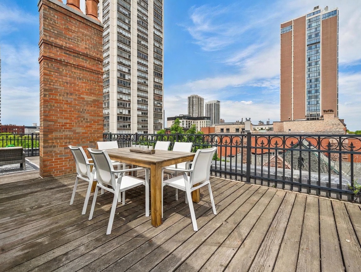 This rooftop deck offers an expansive urban view, featuring a wooden dining set centered on a weathered wood-plank floor. A prominent brick chimney structure stands to the left, while a black metal railing encloses the space, providing a clear vantage point of the surrounding high-rise buildings. The scene is captured from a slightly elevated perspective, emphasizing the open-air atmosphere and the city skyline backdrop.