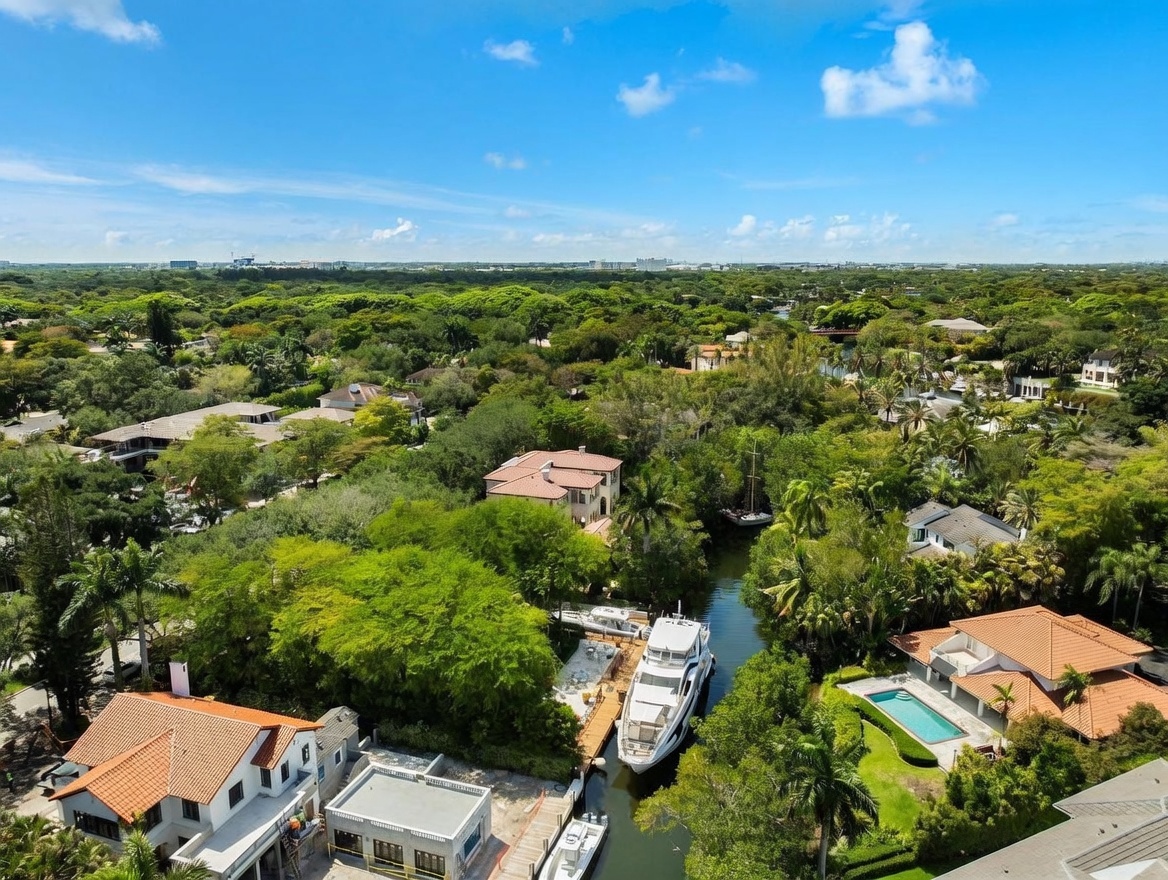 This high-angle aerial view captures a lush, tropical residential waterfront property nestled among dense greenery. A large white yacht is docked in a narrow canal that winds through the landscape, flanked by multi-story homes with terracotta-tiled roofs and a private swimming pool. The perspective provides a comprehensive look at the expansive, tree-filled neighborhood under a bright, clear blue sky.