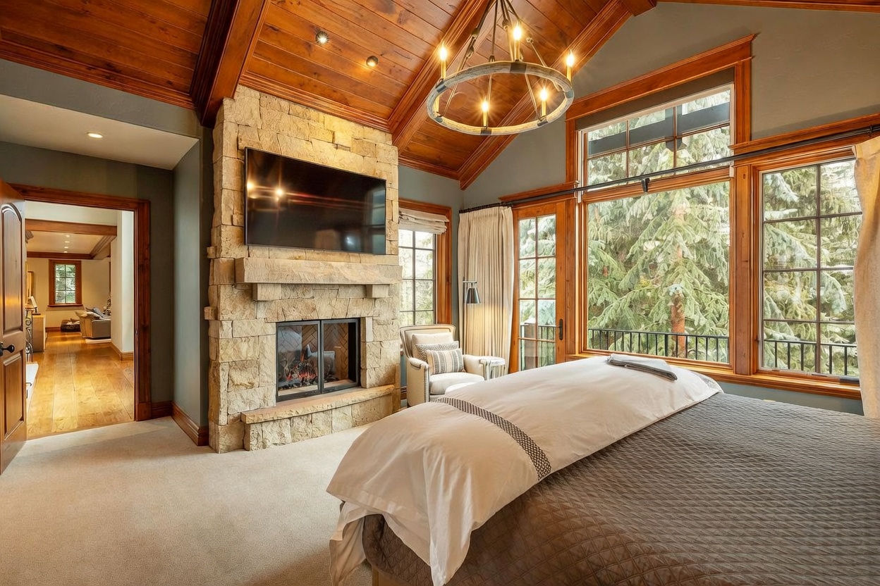 This primary bedroom features a vaulted wood-paneled ceiling and a prominent stone-clad chimney breast housing a television and fireplace. Large windows provide expansive views of the surrounding trees, while a neutral-toned bed with a quilted coverlet serves as the focal point. The room is connected to an adjacent hallway, showcasing a cohesive rustic-luxe aesthetic with warm wood tones and soft textures.