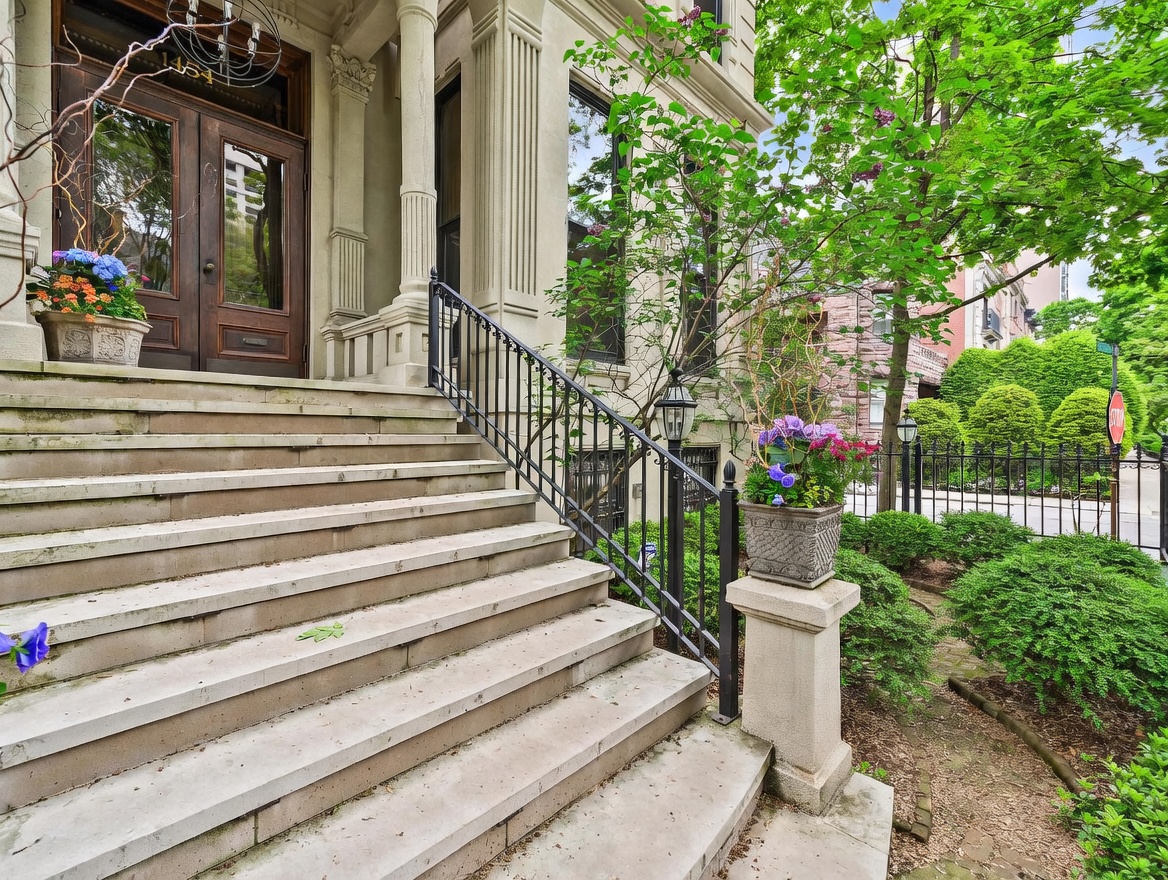 This elegant entryway features a wide set of stone steps leading up to a grand double-door entrance framed by classical columns. The facade is adorned with decorative planters filled with vibrant flowers, and a black wrought-iron railing provides a sophisticated touch. The perspective is from the base of the stairs, looking up toward the entrance, capturing the stately architectural details and the lush greenery of the surrounding neighborhood.