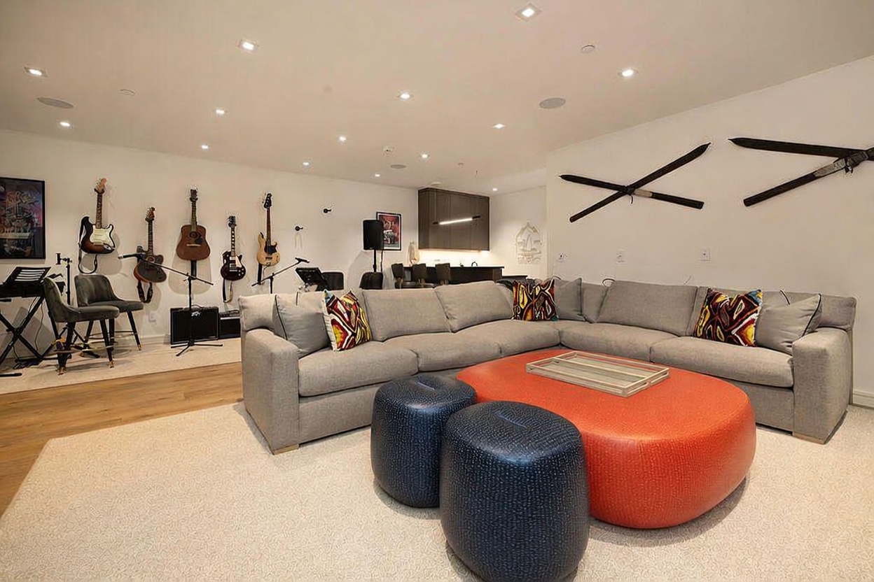 This spacious, contemporary living room features a large, light-gray sectional sofa accented with colorful patterned throw pillows and a prominent orange ottoman. The room is designed as a multi-functional entertainment space, showcasing a collection of guitars mounted on the wall and a bar area in the background. Recessed lighting illuminates the neutral-toned space, which is decorated with wall-mounted skis and framed artwork, creating a relaxed and inviting atmosphere.