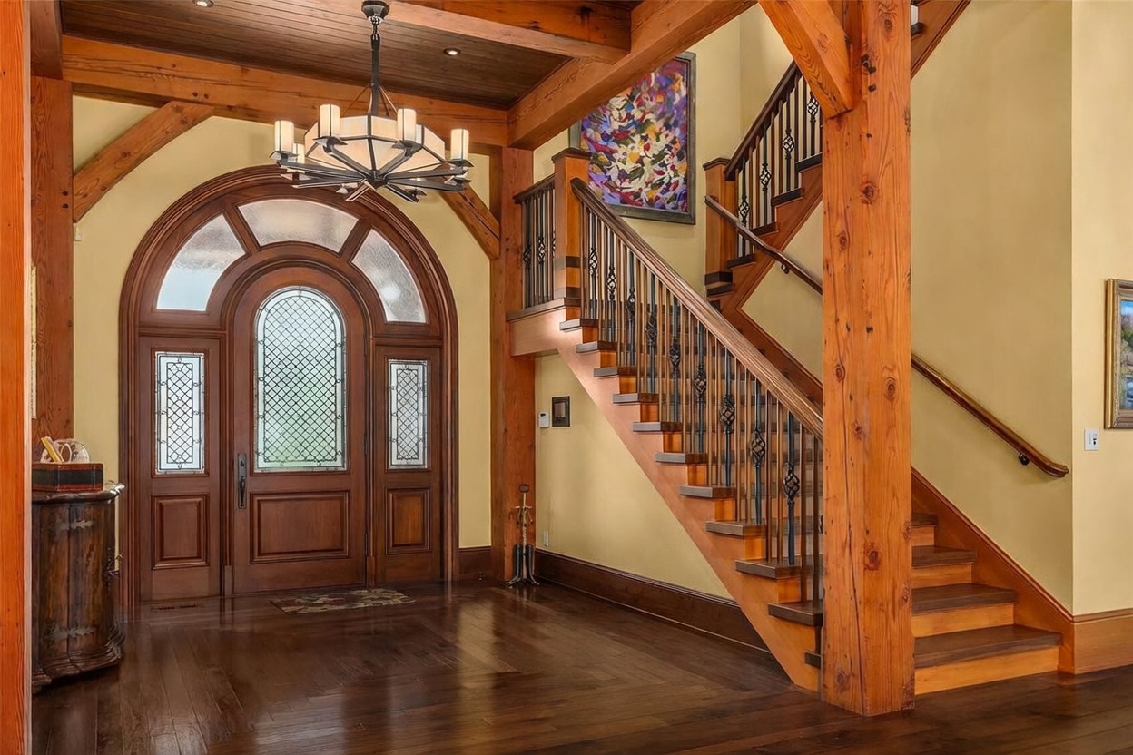 This grand entryway features a striking combination of rustic timber framing and elegant craftsmanship, highlighted by a large arched wooden front door with decorative glass panels. A prominent staircase with dark metal railings and wood treads ascends to the upper level, while a large, colorful abstract painting adds a vibrant focal point to the wall. The space is illuminated by a sophisticated multi-armed chandelier suspended from a wood-paneled ceiling, creating a warm and inviting atmosphere.
