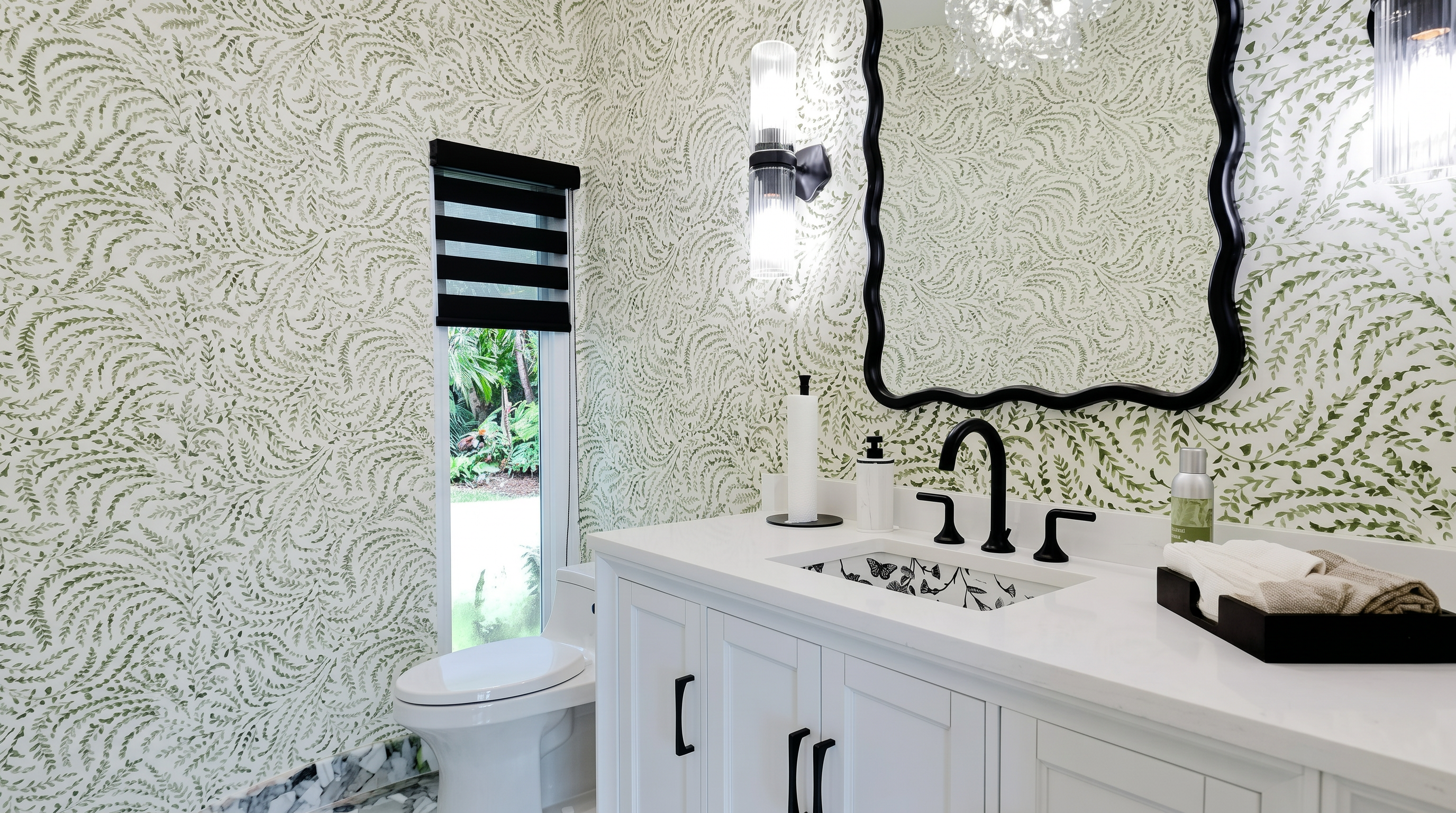 This bathroom features a striking vanity with a white countertop and a decorative sink bowl, complemented by a scalloped, modern black mirror. The walls are adorned with an intricate, olive-green fern-patterned wallpaper that adds a nature-inspired, high-style touch to the room. A sleek wall sconce and a high-contrast window blind enhance the contemporary and elegant aesthetic of this powder room.