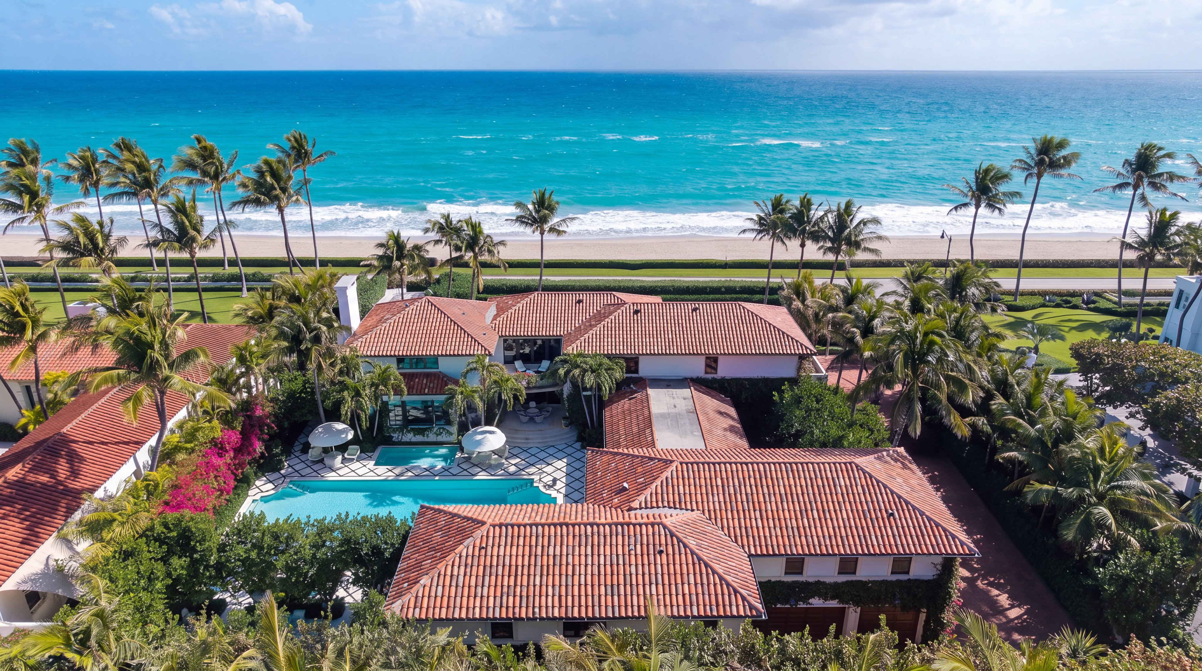 This high-angle aerial view captures a luxurious beachfront estate featuring a classic Mediterranean-style architecture with a terracotta tile roof. The property includes a private swimming pool set within a landscaped courtyard, surrounded by lush tropical greenery and palm trees. The home is situated directly across from a pristine sandy beach and the turquoise waters of the ocean, offering a prime coastal location.