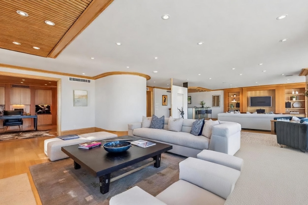 This expansive, open-concept living area features a sophisticated neutral palette with light-toned sofas and a large, dark-finished coffee table as a central focal point. The room is defined by a curved wall, a slatted wood ceiling detail, and integrated shelving units that house a television and decorative items. The perspective captures a spacious, airy layout that flows seamlessly into an adjacent office space and a bar area, creating an ideal environment for entertaining.
