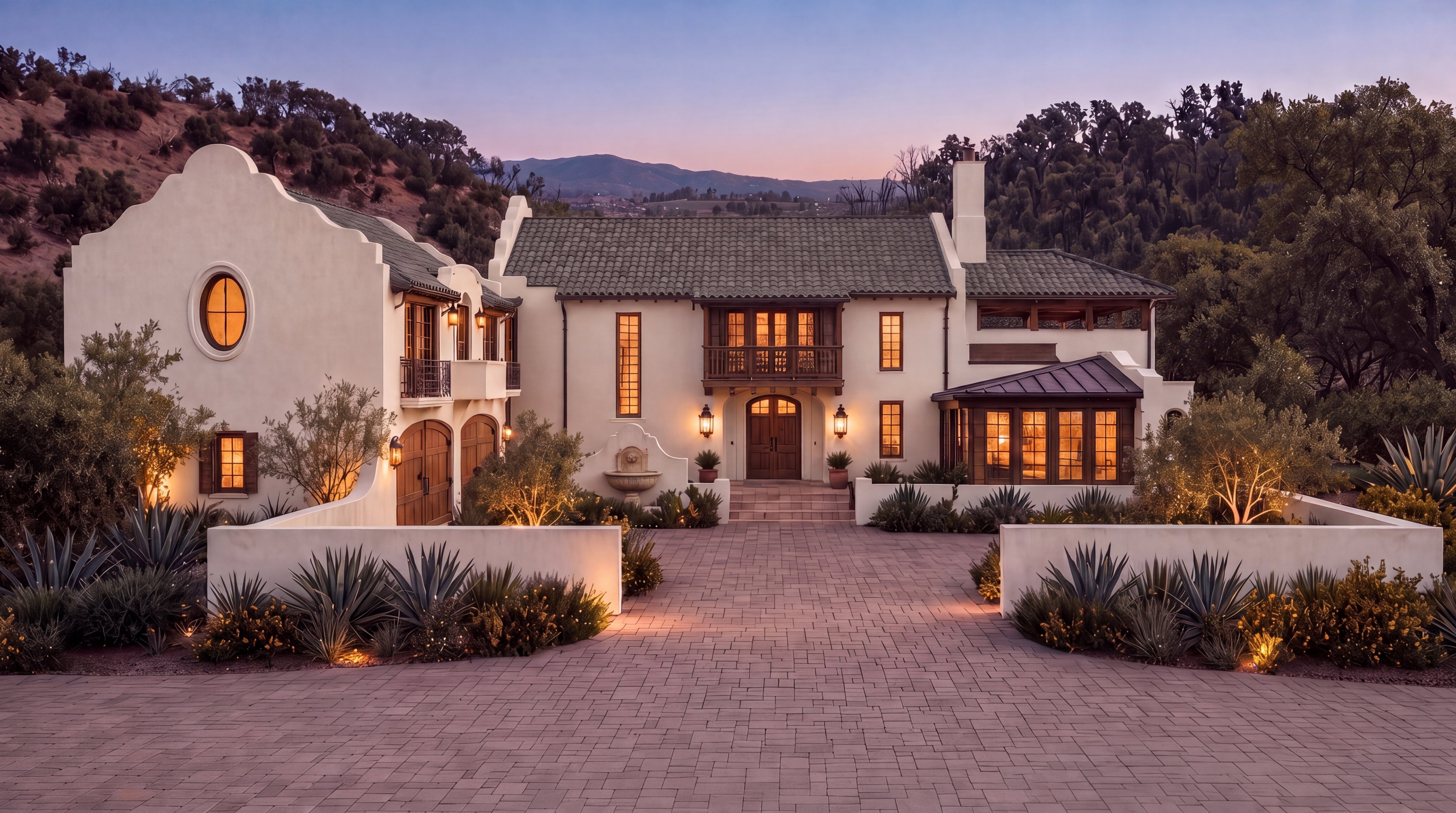 This elegant Spanish Colonial-style estate features a grand, symmetrical facade with light stucco walls, green tiled roofing, and rich wooden accents. A spacious, paved paver driveway leads to the impressive double-door entrance, complemented by warm exterior lighting and lush desert landscaping including agaves. The home creates a luxurious and inviting atmosphere, perfectly nestled against a scenic hilly backdrop at twilight.