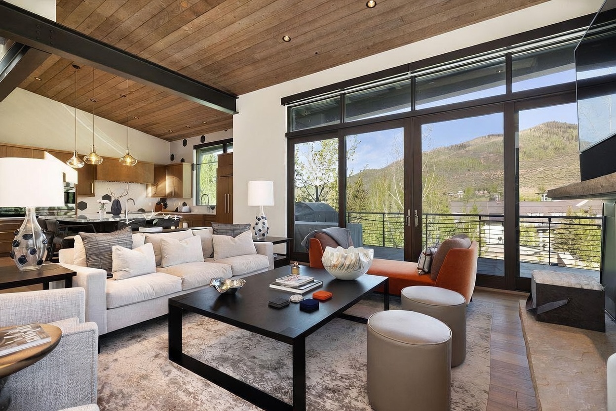 This open-concept living area features a contemporary design with a warm wood-paneled ceiling and a neutral color palette. The space is anchored by a large, dark coffee table surrounded by a plush sofa and orange accent seating, with a stone-clad chimney breast visible on the right. Large glass doors provide an expansive view of the mountain landscape, filling the room with natural light.