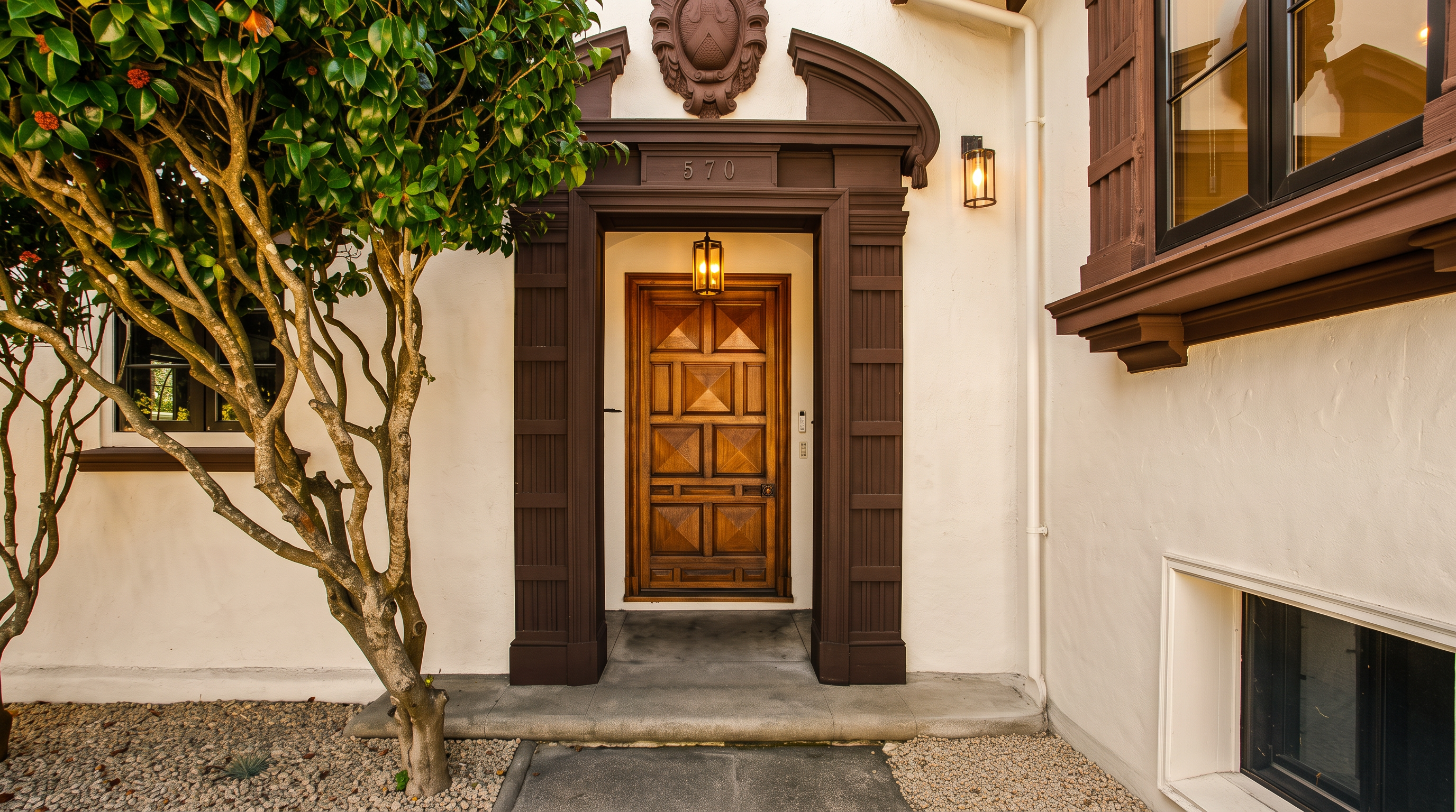 This inviting entryway features a classic wooden door with a geometric panel design, framed by a dark, ornate architectural surround. A decorative crest sits above the doorway, while a hanging lantern provides warm illumination. The scene is framed by a mature tree on the left and a small, manicured shrub on the right, creating a welcoming and established curb appeal.