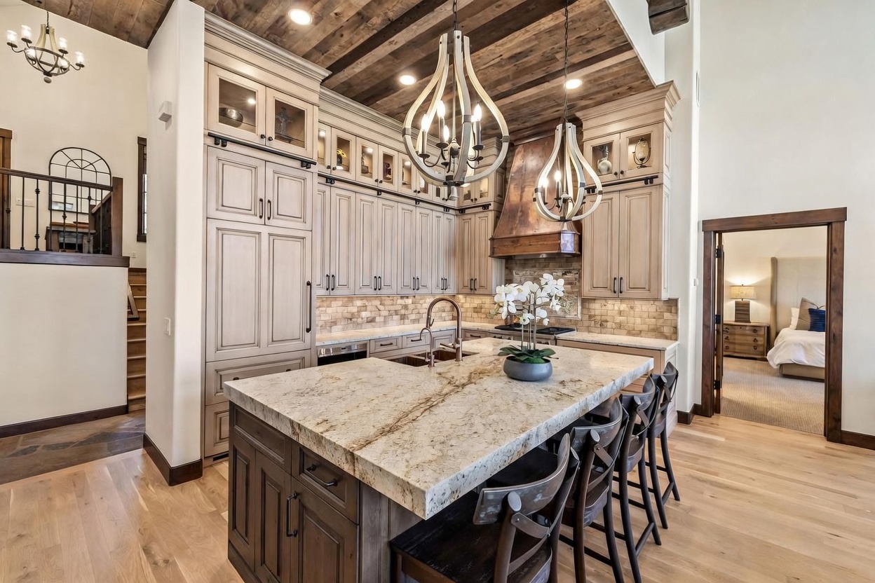 This rustic-inspired kitchen features a large central island with a stone countertop and seating, complemented by light-toned cabinetry and a distinctive wood-plank ceiling. Two decorative pendant lights hang above the island, while a copper-toned range hood serves as a focal point over the stove area. The space flows seamlessly into an adjacent room, with light wood flooring and a staircase visible to the left, creating an open and inviting atmosphere.
