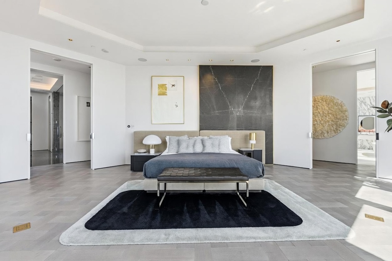 This spacious primary bedroom features a central bed with a neutral upholstered headboard set against a dramatic, dark stone-clad wall. The room is anchored by a layered rug with a bold black center and is flanked by two doorways leading to adjacent spaces, including a glimpse of a bright, marble-finished bathroom. The minimalist aesthetic is complemented by light-toned wood flooring and a low-profile bench at the foot of the bed, creating a serene and sophisticated atmosphere.