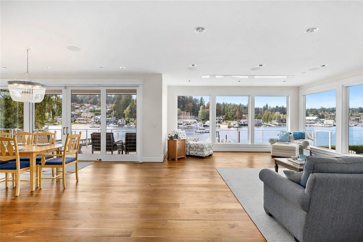 This bright, open-concept living and dining area features light wood flooring and expansive windows that provide panoramic views of the waterfront and marina. The space is furnished with a comfortable grey sofa, a patterned accent chair, and a wooden dining set illuminated by a crystal chandelier. The neutral color palette and abundant natural light create an airy, inviting atmosphere perfect for waterfront living.