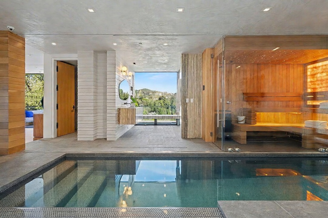 This luxurious indoor spa area features a sleek, tiled swimming pool in the foreground and a glass-enclosed sauna with wood-paneled walls and integrated lighting. The space is finished with neutral stone flooring and light wood accents, leading to a vanity area with a circular mirror and a large window that frames a scenic outdoor view. The design emphasizes a serene, high-end wellness atmosphere with clean lines and a seamless transition between the water feature and the living space.
