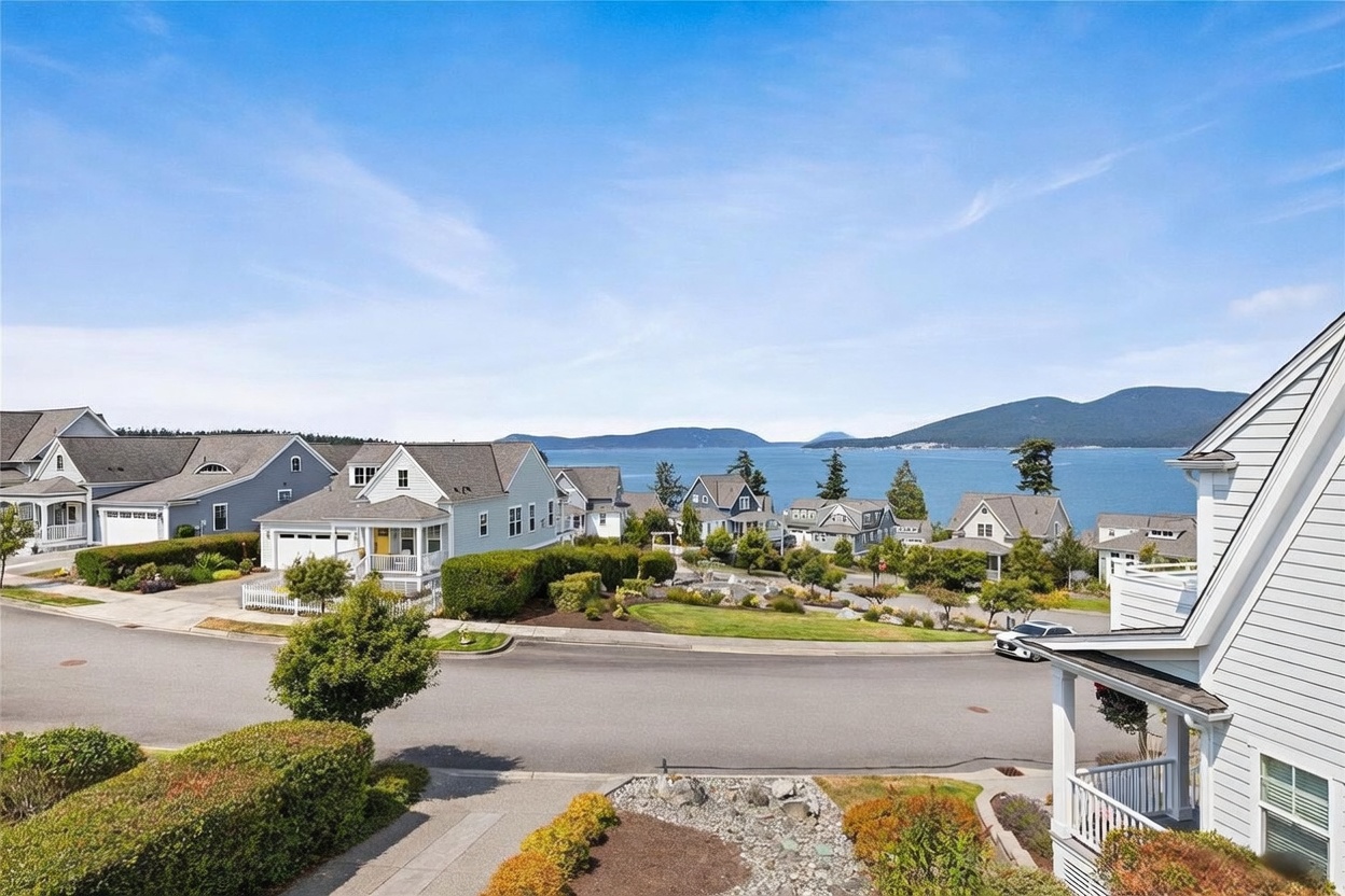 This elevated perspective captures a picturesque coastal neighborhood featuring charming, light-colored homes with classic architectural details. The scene overlooks a quiet residential street that curves toward a stunning, expansive view of the deep blue bay and distant mountains under a clear, bright sky. Lush green landscaping, manicured hedges, and a rocky garden foreground frame the tranquil, scenic setting.