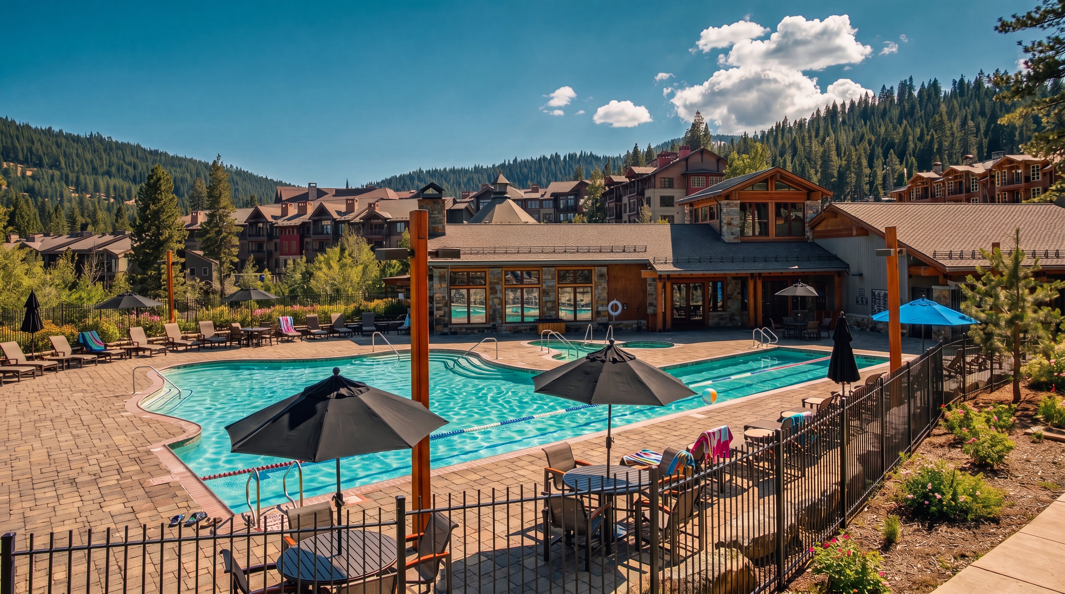 This image features an inviting outdoor swimming pool and spa complex situated within a resort-style community, framed by lush pine forests and mountain slopes. The area includes a stone-accented clubhouse, ample patio space with numerous lounge chairs and tables, and a secure metal perimeter fence. It offers a peaceful and well-maintained leisure space ideal for residents or guests.