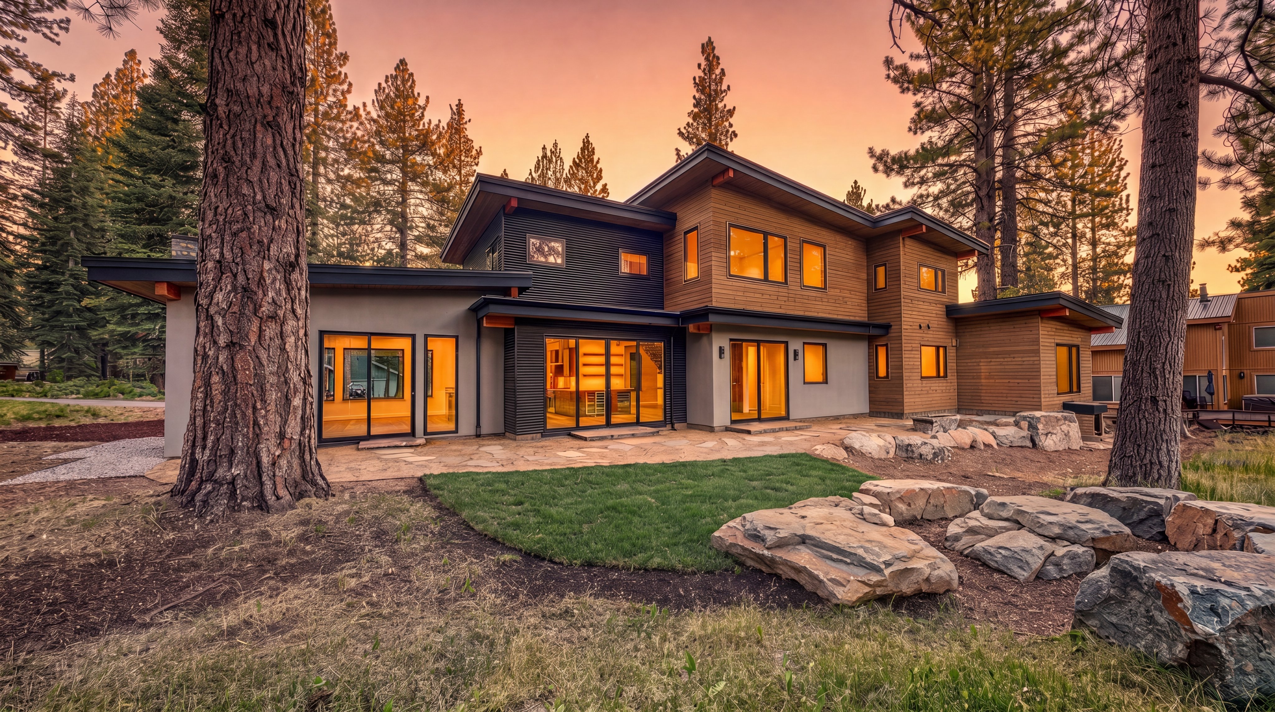 This contemporary mountain home features a striking mix of horizontal wood siding, dark corrugated metal, and smooth gray stucco. The rear facade showcases large glass sliding doors that open onto a stone patio, seamlessly blending indoor and outdoor living spaces. Surrounded by tall pine trees, the residence offers a serene, modern aesthetic with a multi-level roofline and warm interior lighting visible through the windows.