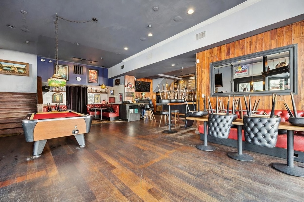 This image captures the interior of a commercial bar or lounge space, featuring a pool table in the foreground and a mix of booth and high-top seating. The room is characterized by dark wood flooring, a deep blue ceiling, and a prominent wood-paneled wall with a large mirror. The perspective is wide-angle, showcasing the layout of the establishment with chairs stacked on tables, suggesting the space is currently closed or being prepared.