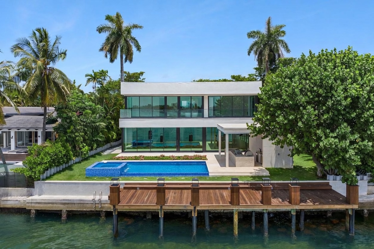 This modern waterfront residence features a sleek, minimalist design with expansive floor-to-ceiling glass walls on both levels, offering seamless indoor-outdoor connectivity. The backyard includes a rectangular swimming pool with an integrated spa, a covered outdoor kitchen area, and a private wooden dock extending into the water. Lush tropical landscaping, including tall palm trees, frames the property, creating a serene and luxurious coastal atmosphere.