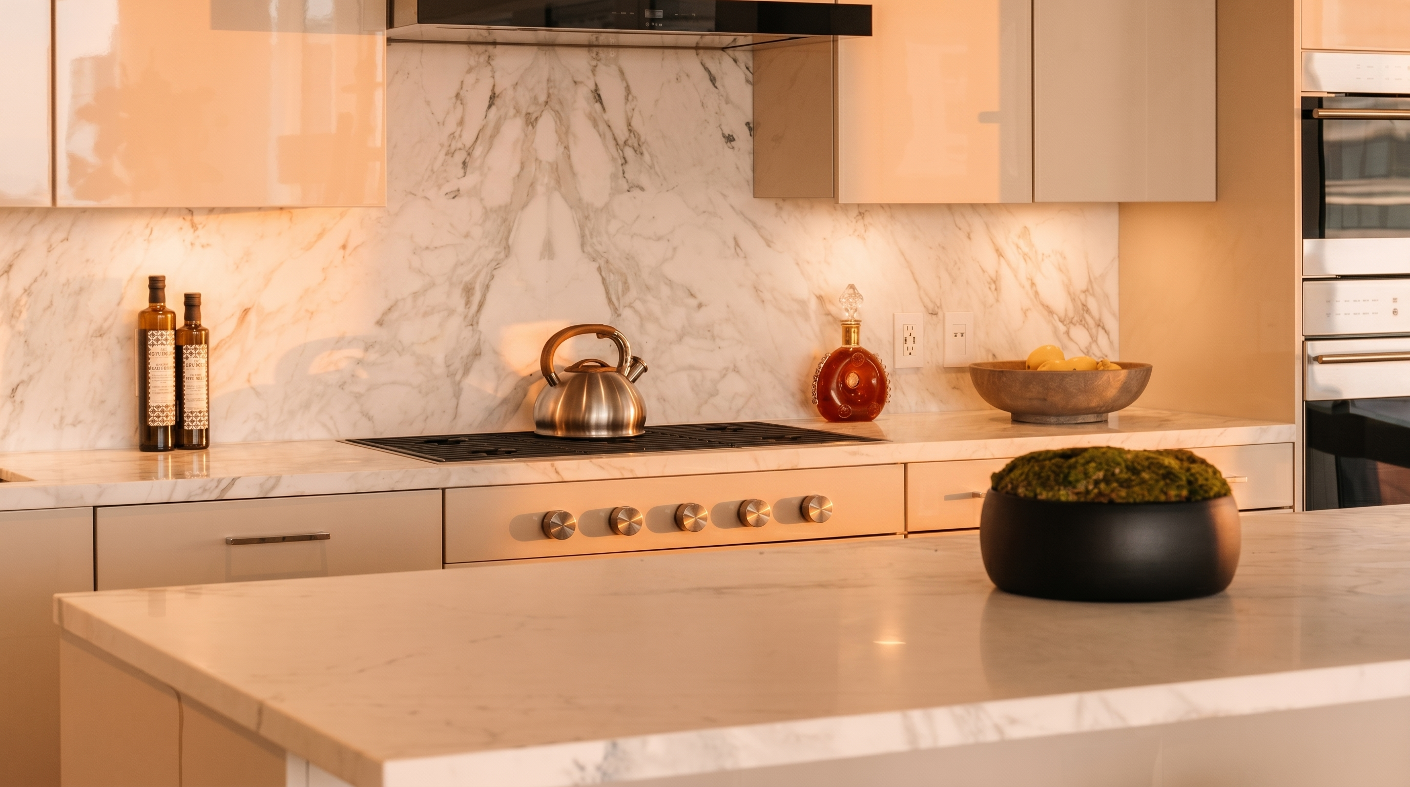 This sleek, modern kitchen features high-end finishes, including light-toned cabinetry and marble countertops and backsplashes. Key elements include a built-in Wolf cooktop, integrated stainless steel ovens, and a spacious center island topped with a decorative moss centerpiece. The neutral color palette and clean lines provide a sophisticated and highly luxurious aesthetic perfect for high-end residential listings.