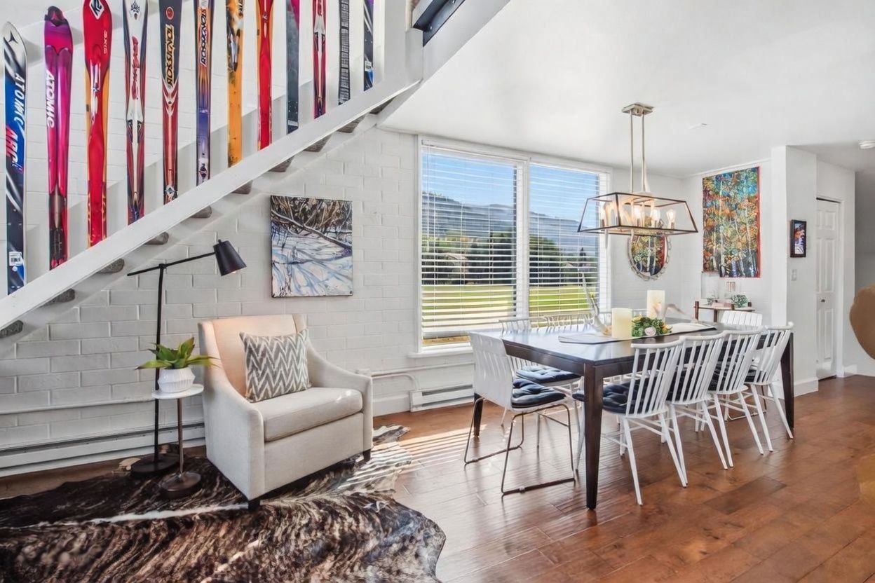 This bright dining area features a dark wood table surrounded by white spindle-back chairs, set against a backdrop of white-painted brick walls. A unique staircase design incorporates vintage skis as decorative balusters, adding a playful, mountain-inspired aesthetic to the space. Large windows provide ample natural light and a scenic view of the outdoors, while a cowhide rug and modern lighting fixture complete the cozy, rustic-chic atmosphere.