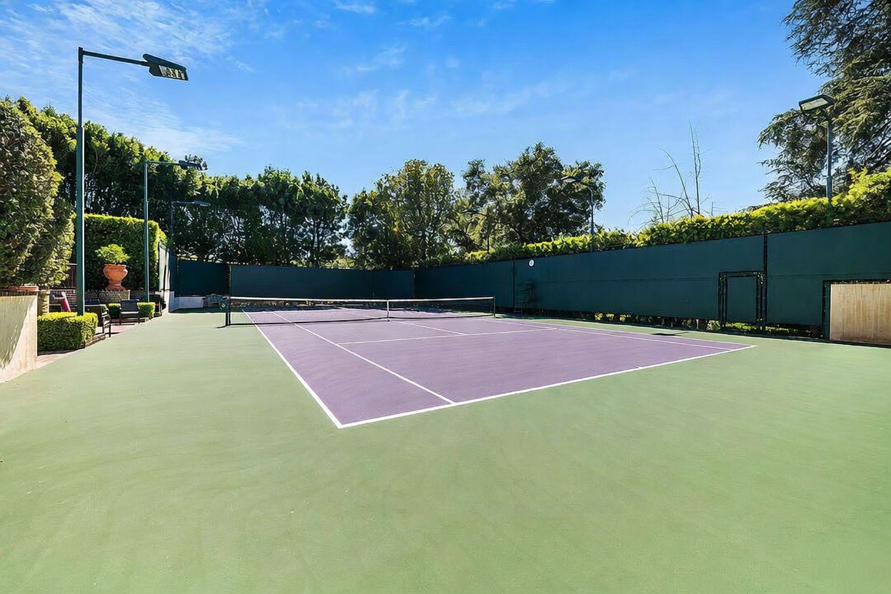 This image showcases a private, well-maintained tennis court featuring a purple playing surface surrounded by a green perimeter. The court is enclosed by tall, dark green privacy fencing and is bordered by lush, mature trees that provide a sense of seclusion. The perspective is from the side of the court, capturing the full length of the playing area under a clear blue sky.