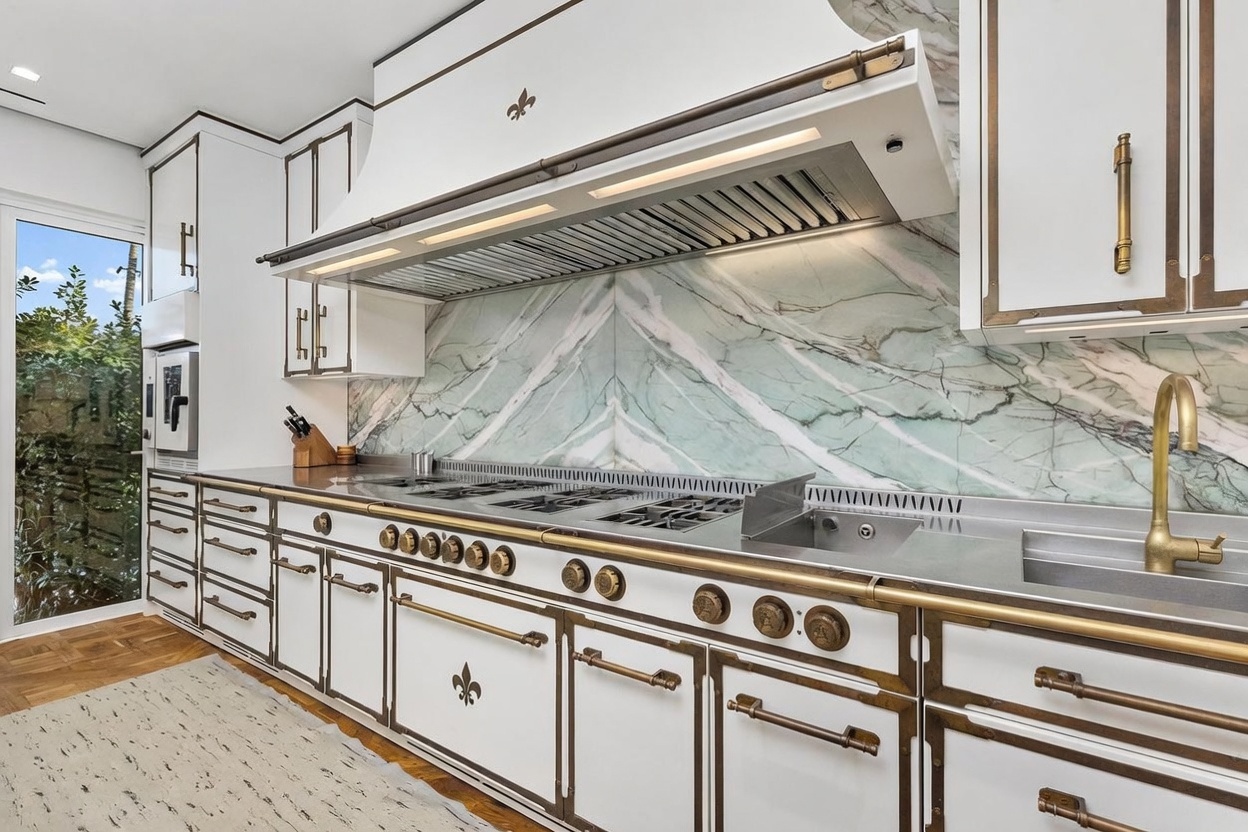 This high-end kitchen features a professional-grade range with a prominent custom hood and a striking green-veined stone backsplash. The cabinetry is finished in white with ornate brass-toned hardware and trim, creating a sophisticated, classic aesthetic. A large window to the left provides natural light, while the integrated sink and expansive counter space emphasize both luxury and functionality.