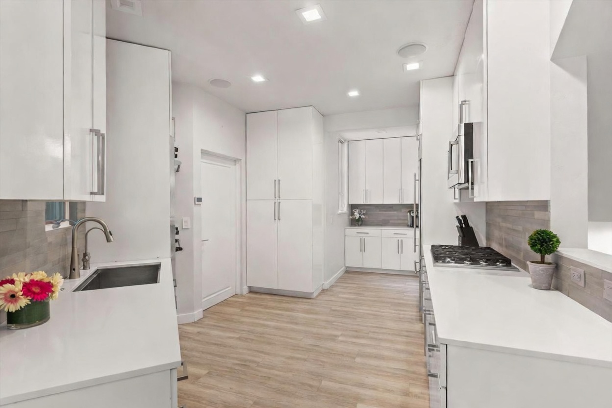 This modern, minimalist kitchen features sleek white cabinetry, light wood-toned flooring, and a clean, bright aesthetic. The space is equipped with a gas cooktop, a built-in microwave, and a stainless steel sink, all complemented by a neutral-toned backsplash. The perspective is a wide-angle shot from the kitchen entrance, showcasing the efficient layout and the seamless integration of appliances into the cabinetry.