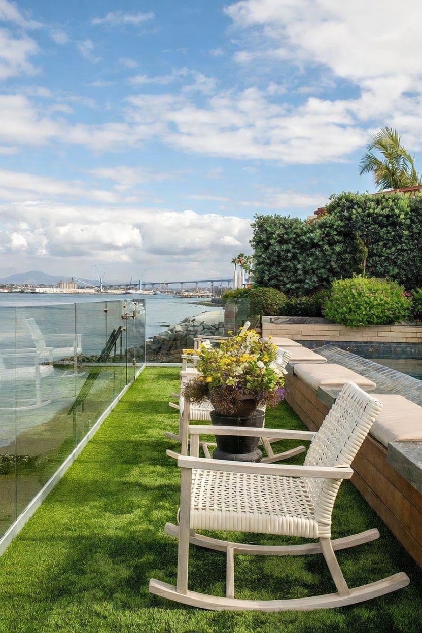 This scenic outdoor patio area features a row of white rocking chairs set on a vibrant green lawn, overlooking a calm bay with a distant bridge. To the right, a built-in stone bench with cushioned seating runs alongside a pool, while a glass railing provides an unobstructed view of the water. The setting is bright and serene, capturing a luxurious waterfront lifestyle with lush landscaping and clear blue skies.