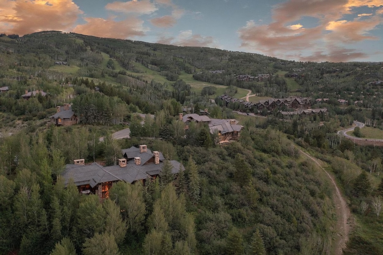 This high-angle aerial view captures a sprawling mountain estate nestled within a dense forest of aspen and pine trees. The home features a complex, multi-gabled roofline with dark shingles and stone exterior walls, blending seamlessly into the rugged, elevated landscape. In the background, a vast mountain slope with ski runs and additional residential clusters stretches toward a dramatic, cloud-filled sunset sky.