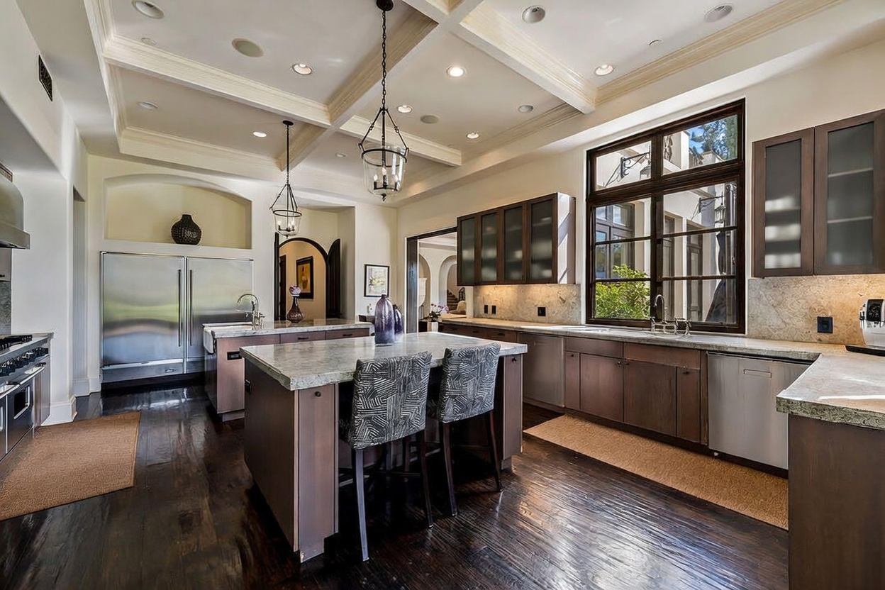 This spacious, high-end kitchen features a coffered ceiling, dark wood flooring, and a large central island with seating for two. The space is equipped with professional-grade stainless steel appliances, including a double-door refrigerator and a range, complemented by stone countertops and dark cabinetry. Large windows provide ample natural light, creating an open and sophisticated atmosphere suitable for both cooking and entertaining.