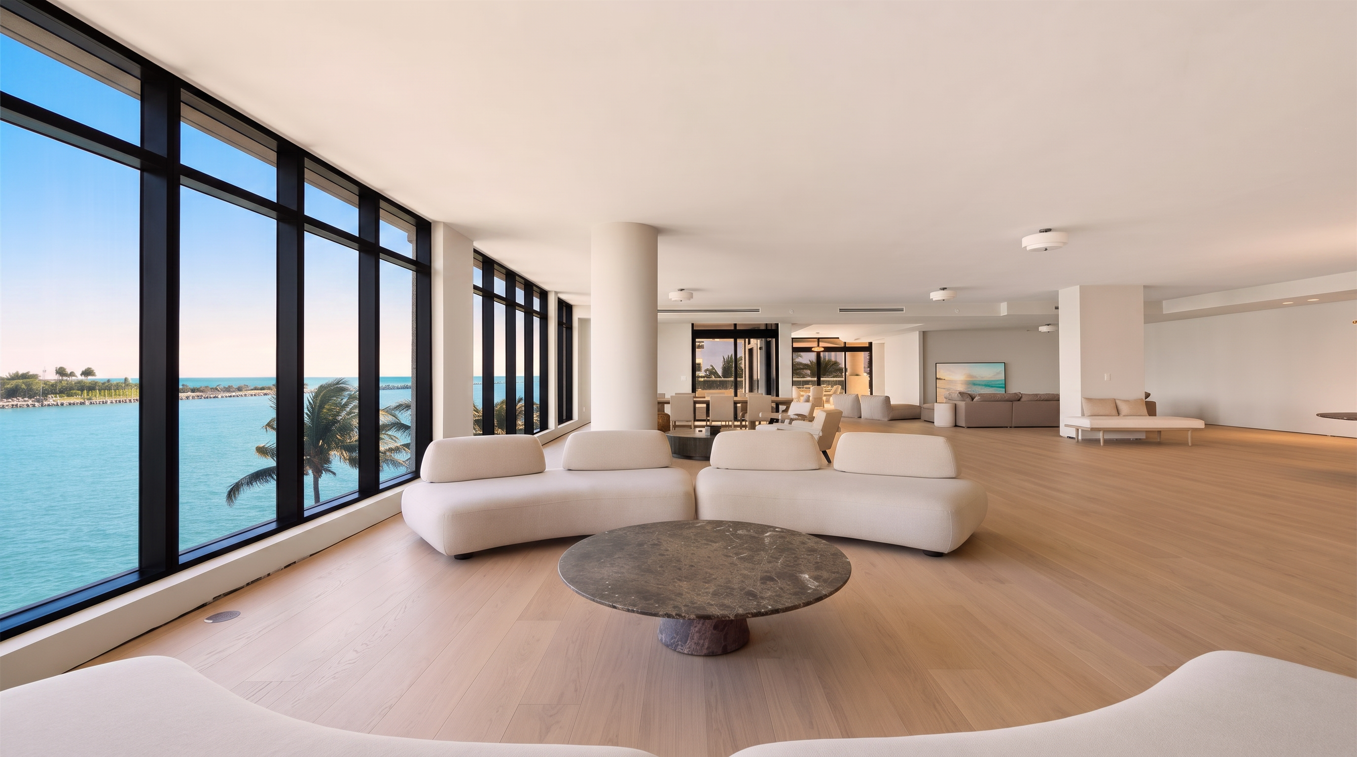 This expansive, open-concept living area features light wood flooring and a neutral color palette that emphasizes the bright, airy atmosphere. A large, circular stone coffee table sits between two curved, cream-colored sofas, while floor-to-ceiling windows provide panoramic views of the ocean and palm trees. The space flows seamlessly into a distant dining area, creating a sophisticated and minimalist aesthetic perfect for luxury coastal living.