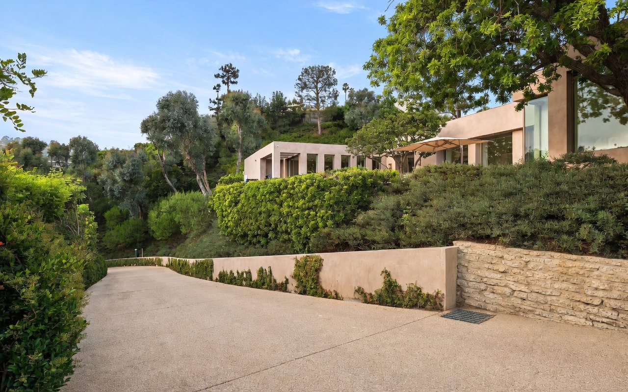This perspective captures a sweeping, paved driveway leading up to a modern, multi-level residence nestled into a lush, hillside landscape. The architecture features clean lines and large glass windows, complemented by mature trees and manicured shrubbery that soften the structure's contemporary aesthetic. The scene conveys a sense of privacy and luxury, with the home appearing integrated into the natural, elevated terrain.