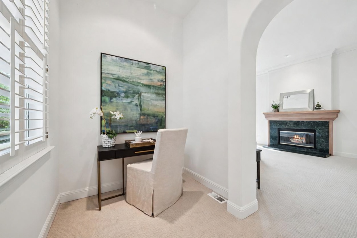 This compact office nook features a sleek dark desk with a beige upholstered chair, accented by a large, colorful abstract painting. The space seamlessly transitions into an adjacent living area through an elegant arched entryway, revealing a fireplace with a dark marble surround. The neutral color palette and clean lines create a sophisticated and inviting home workspace.