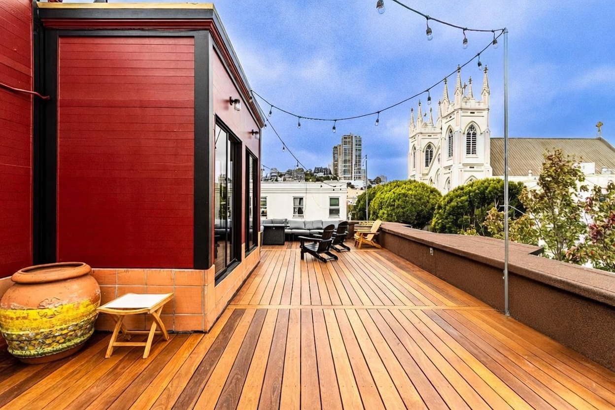 This expansive rooftop deck features warm-toned wood decking and a striking red exterior wall, creating a vibrant outdoor living space. The area is furnished with comfortable seating, including a sofa and chairs, and is illuminated by overhead string lights. A prominent church spire is visible in the background, adding a unique architectural character to the urban skyline view.