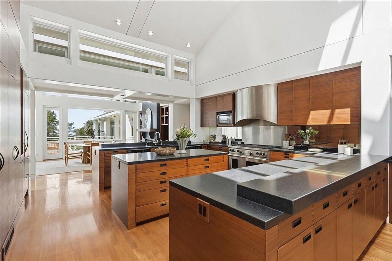 This spacious, modern kitchen features warm-toned wood cabinetry, dark stone countertops, and a large central island that provides ample workspace. The room is defined by its high, vaulted ceiling and a series of clerestory windows that flood the area with natural light, creating an airy and open atmosphere. A view through the adjacent dining area reveals a bright, outdoor deck, emphasizing the home's seamless connection between indoor and outdoor living spaces.