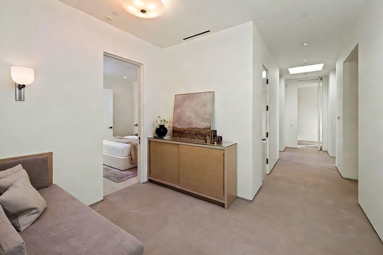 This interior view captures a minimalist hallway and transition space featuring light-toned stone flooring and crisp white walls. A sleek wooden console table sits against the wall, topped with a piece of abstract art, while a doorway leads into a bedroom visible in the background. The perspective is eye-level, emphasizing the clean lines, modern architectural details, and the bright, airy atmosphere of the home.
