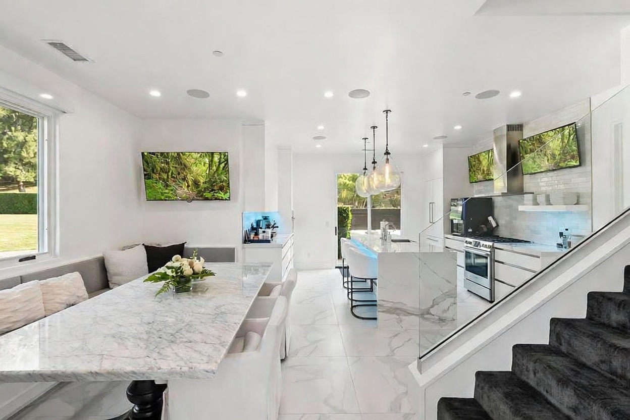 This is a bright and modern kitchen and dining area featuring white marble countertops and flooring. The kitchen includes stainless steel appliances, a central island with bar seating, and pendant lighting. The dining area has a marble-topped table with white chairs and a built-in bench, creating a cohesive and luxurious space.