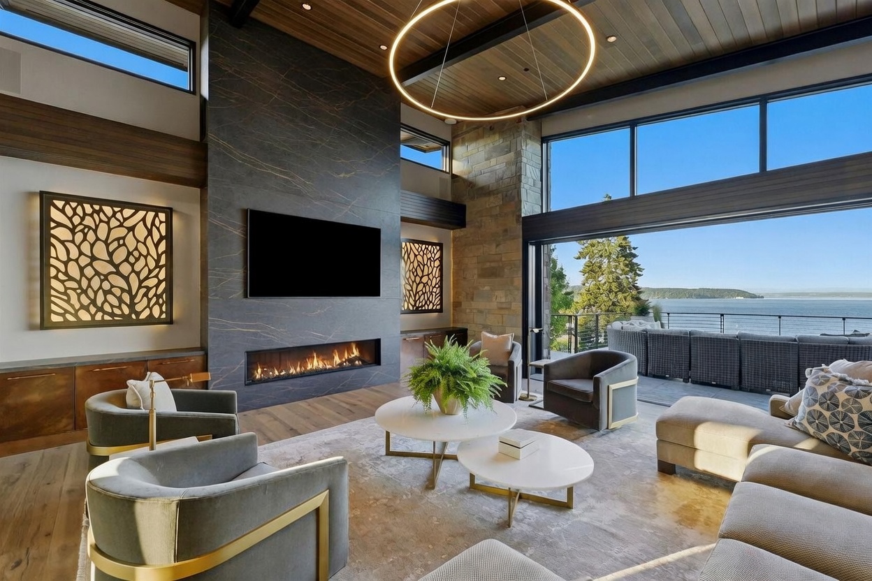 This expansive living room features a striking dark stone-clad chimney breast with a linear fireplace and a mounted television. The space is anchored by a large area rug, two circular coffee tables, and plush seating, all oriented toward a wall of glass that opens to a deck with panoramic water views. A large circular pendant light hangs from the wood-paneled ceiling, and decorative wall panels with organic cutouts add a sophisticated, modern touch to the room.