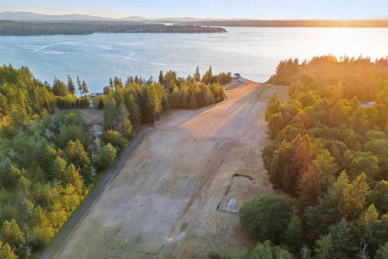 This high-angle aerial view captures a large, expansive plot of land sloping gently toward a scenic waterfront. The property features a wide, open grassy field bordered by dense, mature evergreen forests on either side, with a small concrete foundation visible in the foreground. The perspective highlights the property's proximity to the water and the serene, natural setting during the golden hour.